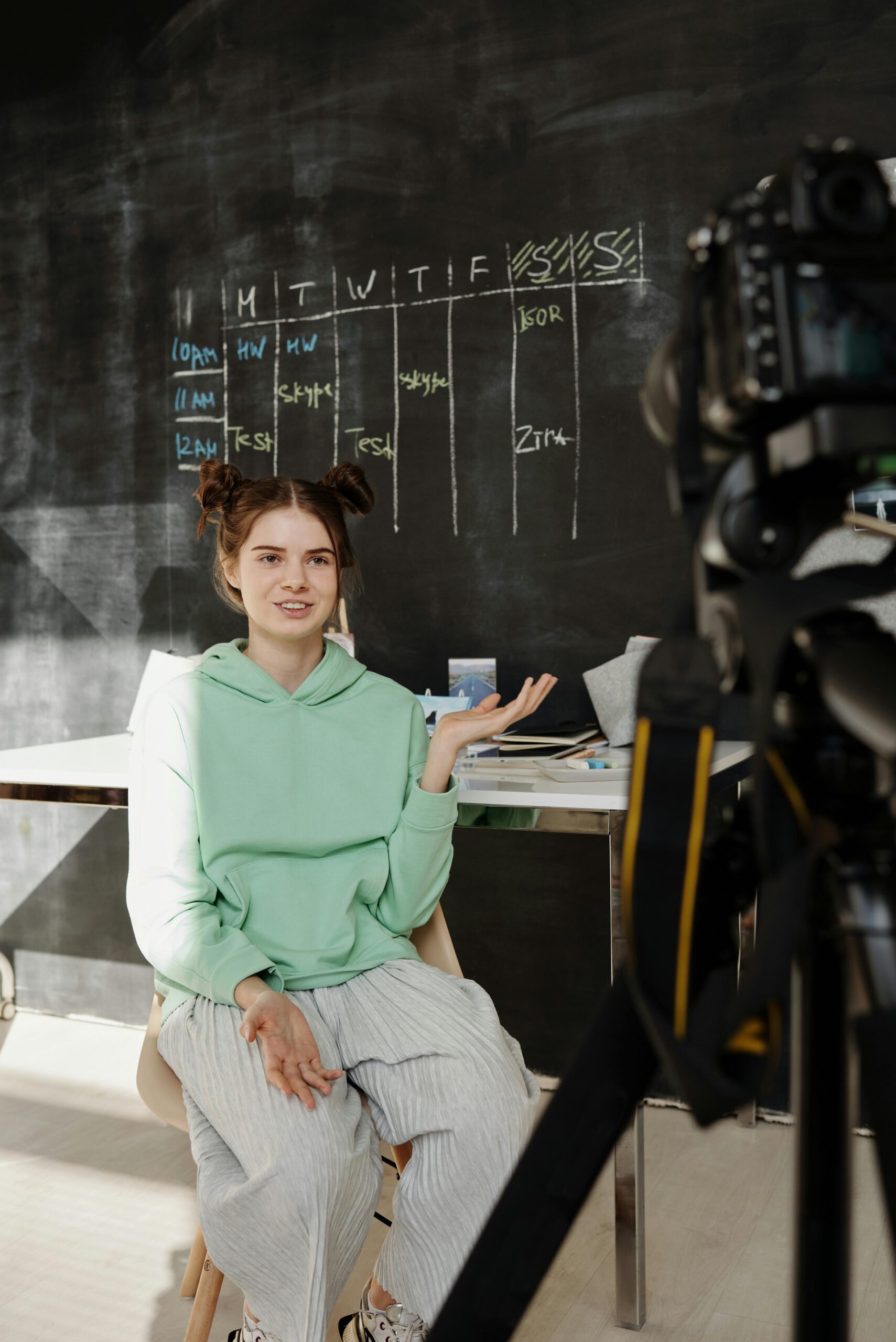 Teenage girl recording a vlog indoors with a camera setup and chalkboard background.